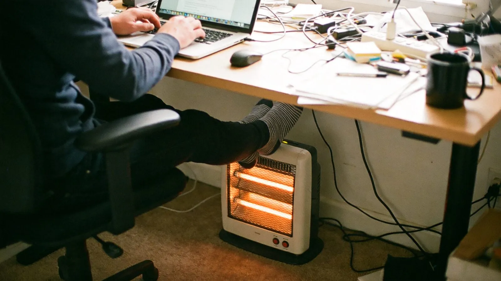 A small quartz personal space heater warming a person's feet under a home office desk while they work on a laptop