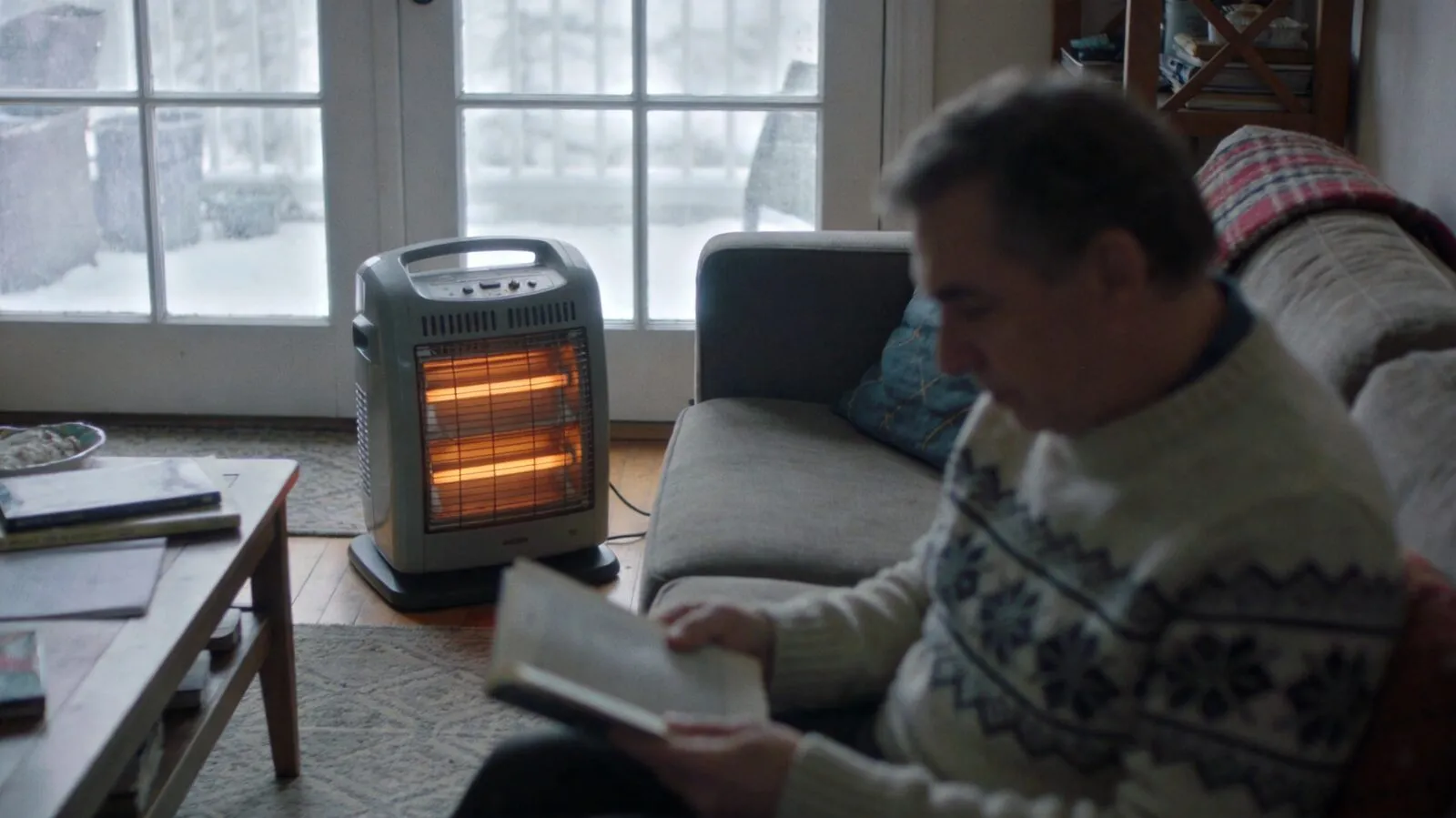 A portable infrared space heater with glowing elements warming a living room while a man reads on the couch on a snowy day